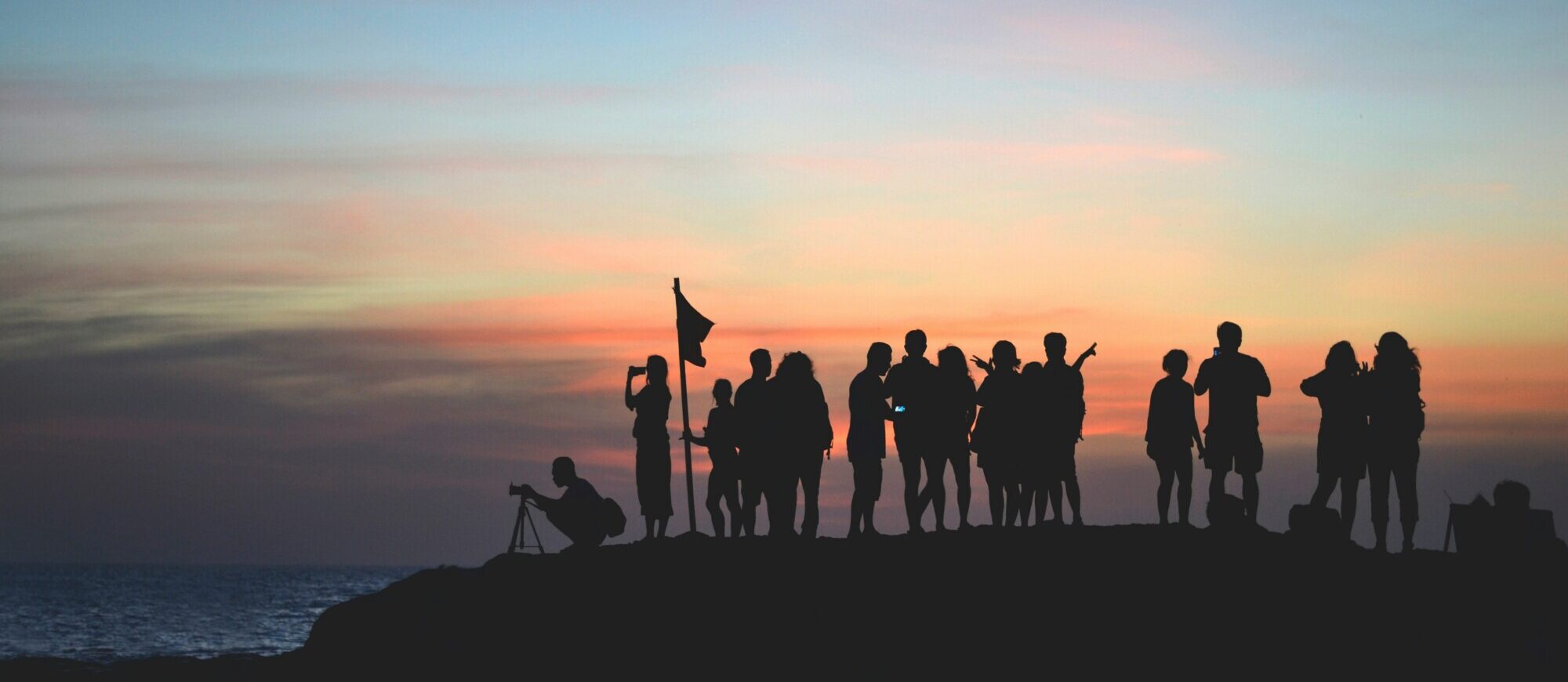 Silhouetten einer Gruppe von Menschen bei Sonnenuntergang. Einige fotografieren oder beobachten den Horizont, der Himmel leuchtet in Rosa-, Orange- und Blautönen.