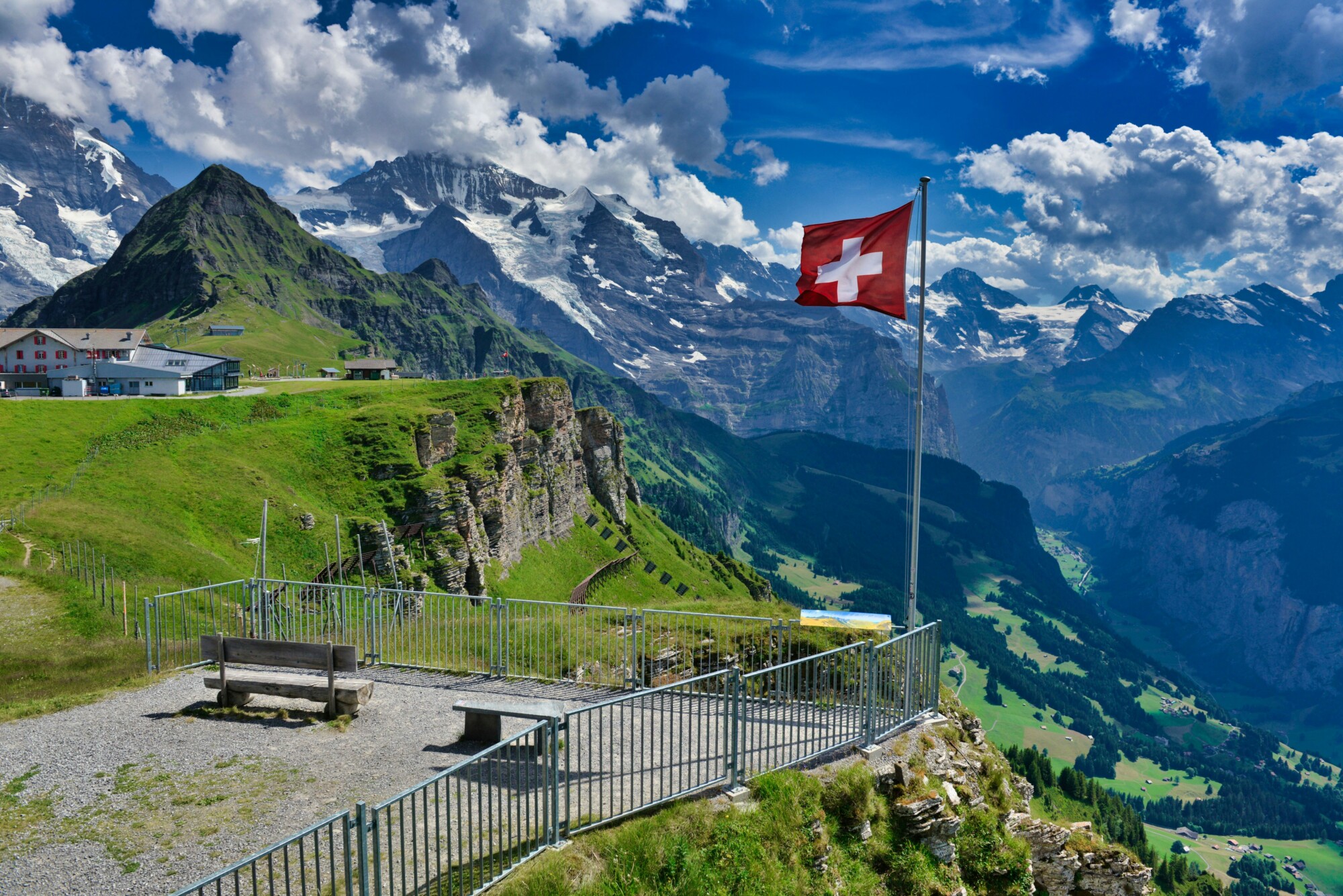 Aussichtspunkt in den Schweizer Alpen mit wehender Schweizer Flagge, grünen Bergwiesen und schneebedeckten Gipfeln unter blauem Himmel.