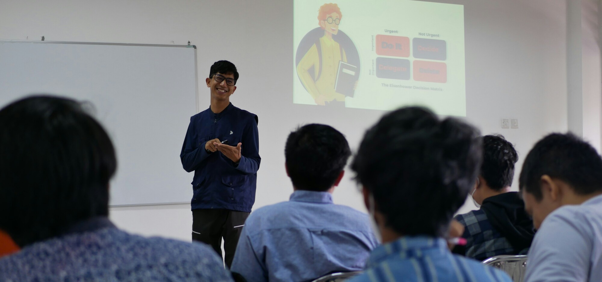 eine Person steht vor einer Gruppe in einem Seminarraum und präsentiert Inhalte, während im Hintergrund eine Folie mit Diagrammen projiziert wird