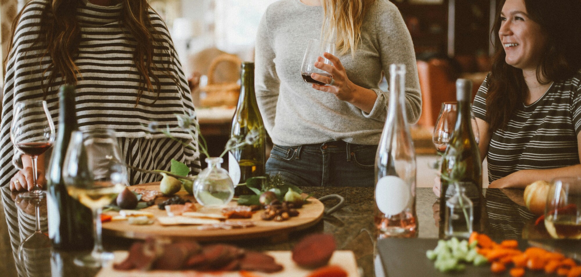 Mehrere Personen stehen in einer Küche zusammen, halten Weingläser in der Hand und unterhalten sich vor einem Tisch mit Weinflaschen und Snacks.