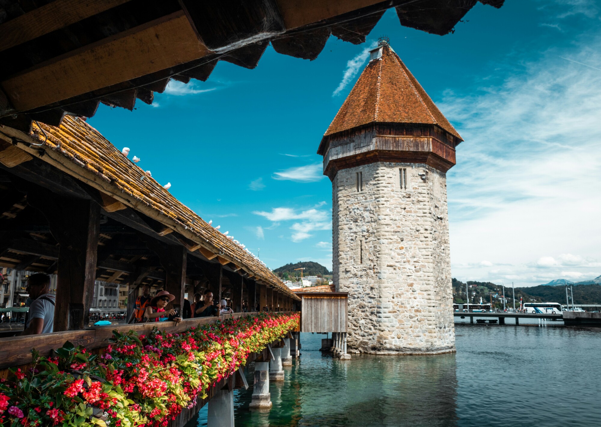 Historische Holzbrücke mit bunten Blumen führt über das Wasser zu einem steinernen Turm, während Menschen die Brücke überqueren und Berge im Hintergrund sichtbar sind.
