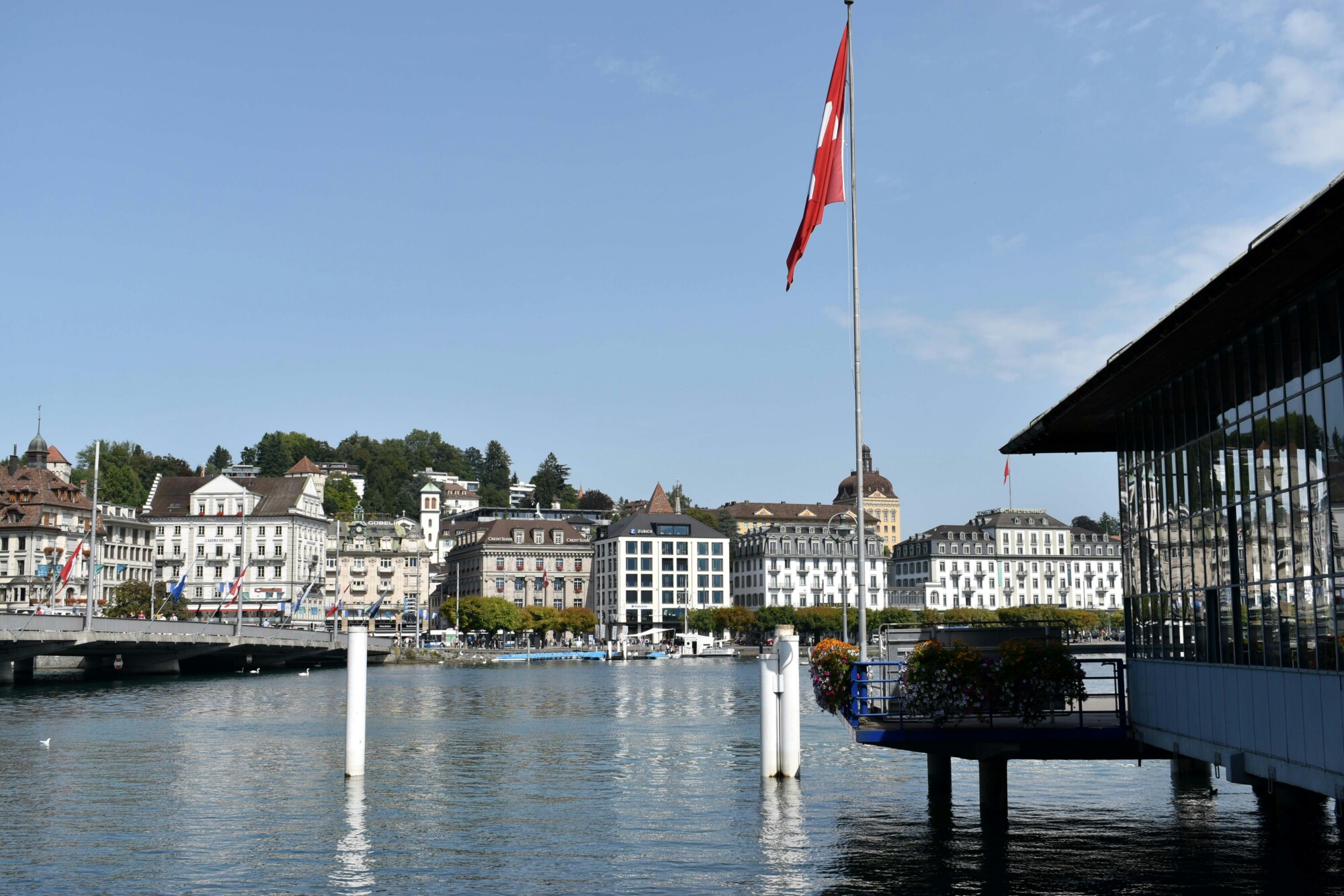 Stadtansicht am Wasser mit Schweizer Flagge auf einem Fahnenmast, historischen Gebäuden entlang des Ufers und einer Brücke im Hintergrund bei klarem Himmel.