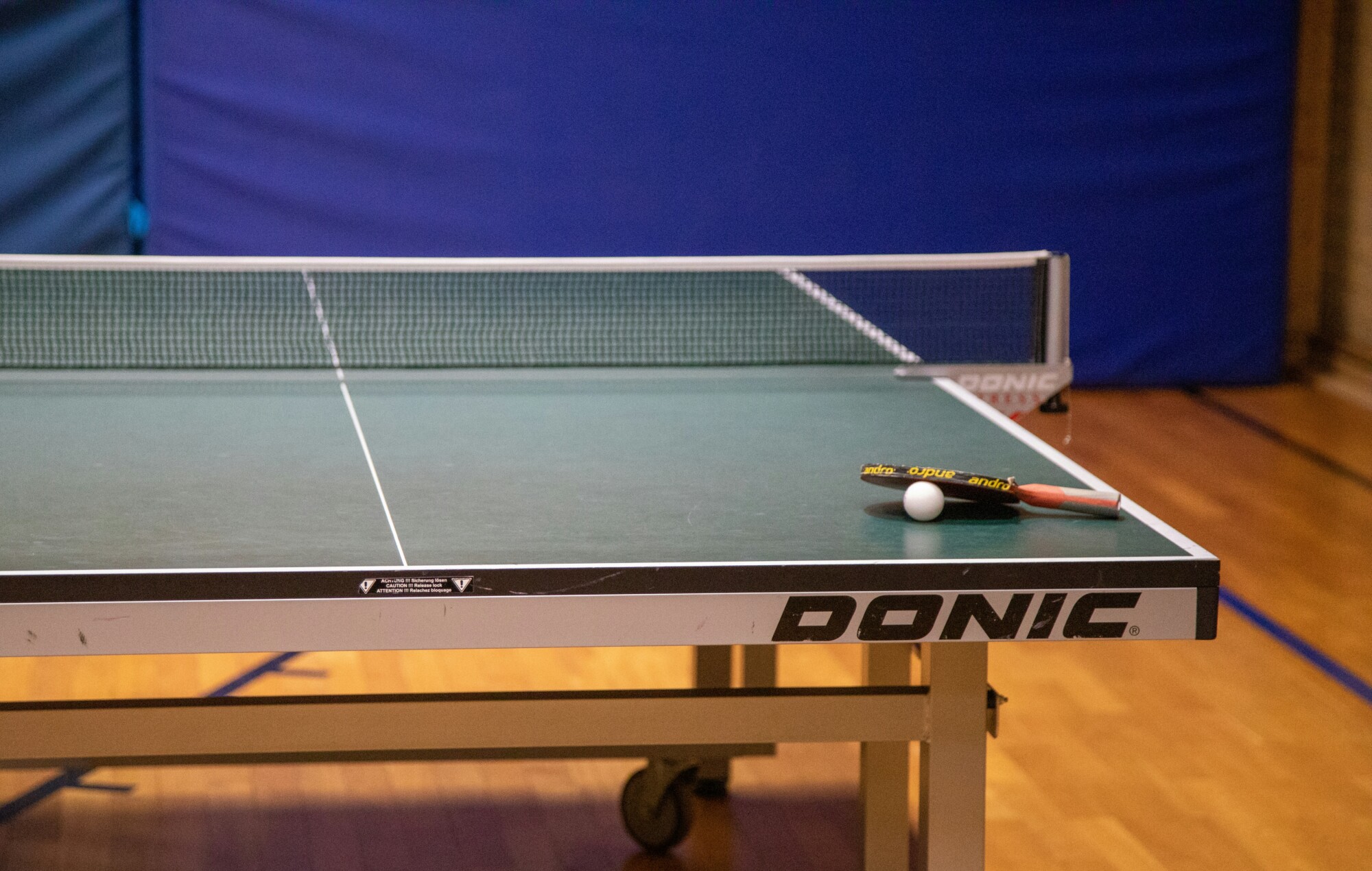 Auf dem Bild ist ein Tischtennistisch in einer Halle zu sehen. Auf dem Tisch liegt ein Schläger und ein Ball.