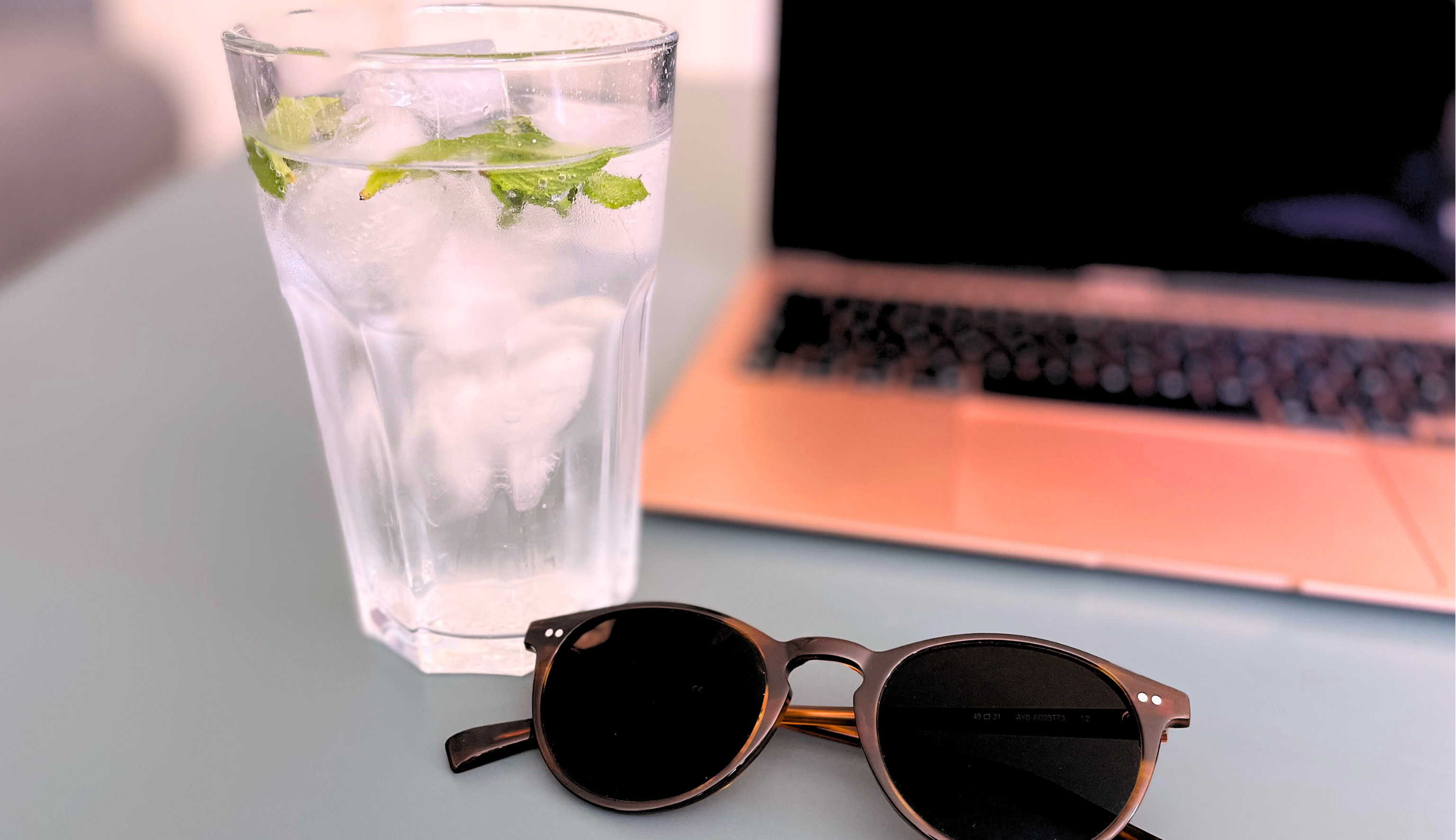 Ein Laptop, eine Sonnenbrille und ein Glas Wasser mit Minze und Eiswürfeln auf einem Tisch