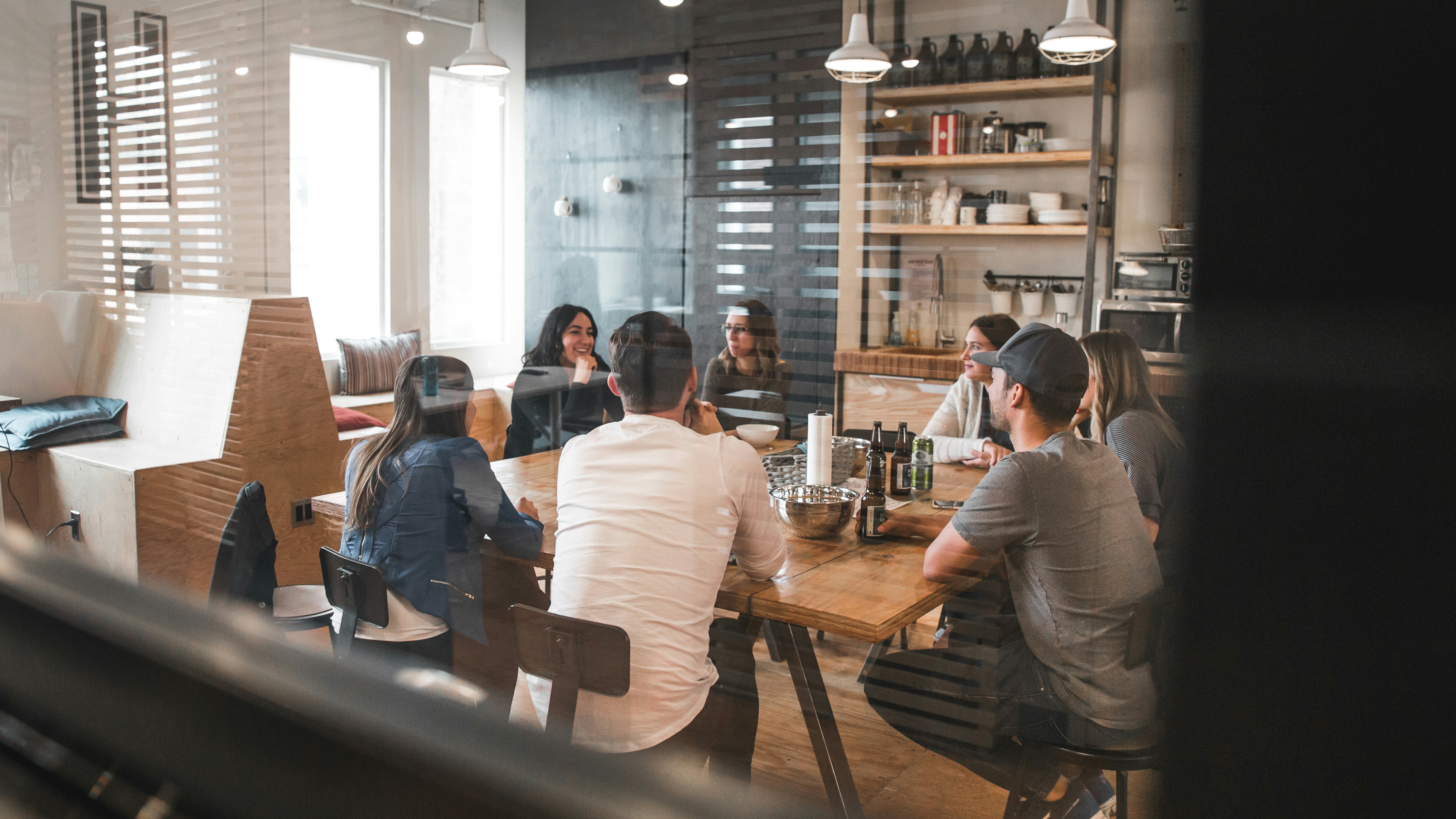13 Tipps wie MitarbeiterInnen wieder gerne ins Büro kommen Ein Team sitzt im Büro bei einem Bier zusammen durch eine Scheibe betrachtet
